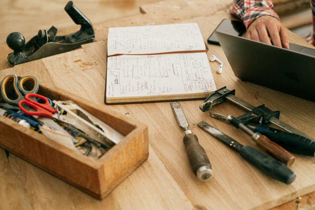 pexels-photo-4491875-4491875 A workspace featuring woodworking tools, a notebook, and a hand on a laptop, symbolizing creativity and craftsmanship.